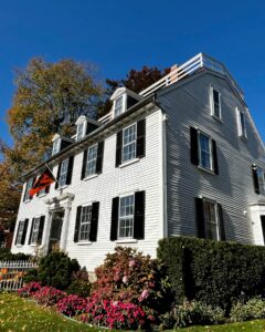 The Ropes Mansion in Salem, Massachusetts, a historic white colonial house with black shutters, a vibrant garden of pink flowers, and trees with autumn leaves under a bright blue sky.
