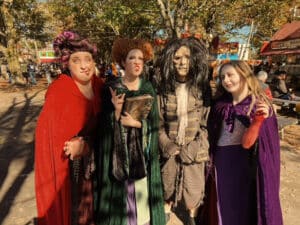 A group of people dressed as the Sanderson Sisters from "Hocus Pocus" and Billy Butcherson standing together in costume at a festive outdoor location in Salem, Massachusetts.