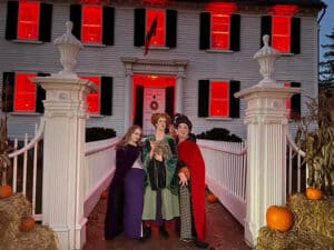 Three people dressed as the Sanderson Sisters from "Hocus Pocus" stand in front of the Ropes Mansion in Salem, Massachusetts. The windows of the mansion are illuminated in red light, creating an eerie and magical atmosphere, while pumpkins and corn stalks decorate the entrance.