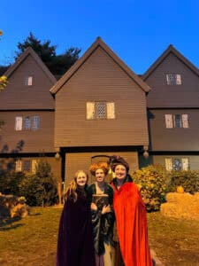 Three people dressed as the Sanderson sisters from Hocus Pocus stand in front of the historic Witch House, posing with smiles. The sky is a deepening blue as the evening approaches, highlighting the unique architecture of the house with its dark wooden exterior.