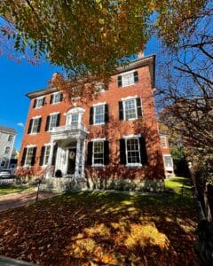 The William Pickman House in Salem, Massachusetts, a red brick Federal-style home with black shutters, a white columned entryway, and autumn leaves covering the front lawn.