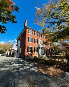 The William Pickman House in Salem, Massachusetts, a historic red brick building with black shutters and a white columned entryway, surrounded by fall foliage.