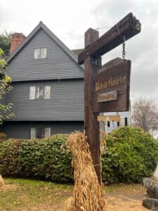The Witch House in Salem, Massachusetts, with its dark wooden exterior visible behind a wooden sign hanging from a post. The sign reads "Witch House," and the yard is decorated with bundles of dried cornstalks, adding to the historic and atmospheric feel of the site.