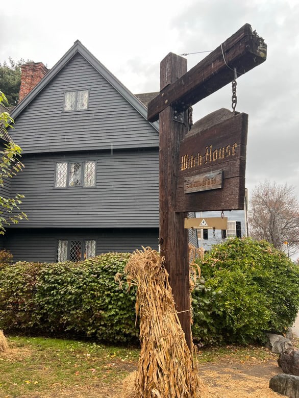 The Witch House in Salem, Massachusetts, with its dark wooden exterior visible behind a wooden sign hanging from a post. The sign reads "Witch House," and the yard is decorated with bundles of dried cornstalks, adding to the historic and atmospheric feel of the site.