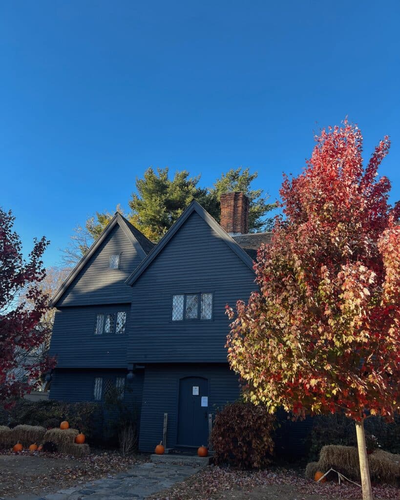 The Witch House in Salem, Massachusetts, is surrounded by colorful autumn trees, pumpkins, and hay bales. The dark exterior of the historic house contrasts with the bright blue sky and vibrant red leaves.