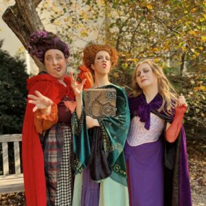 Three women dressed as the Sanderson Sisters from the movie Hocus Pocus in Salem, Massachusetts. The costumes are highly detailed, with colorful capes, dresses, and a spellbook prop.