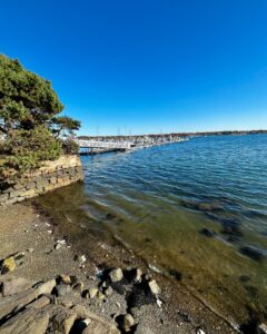 View of the waterfront near the House of the Seven Gables in Salem, Massachusetts, showcasing a stone wall, calm waters, and a marina in the distance.