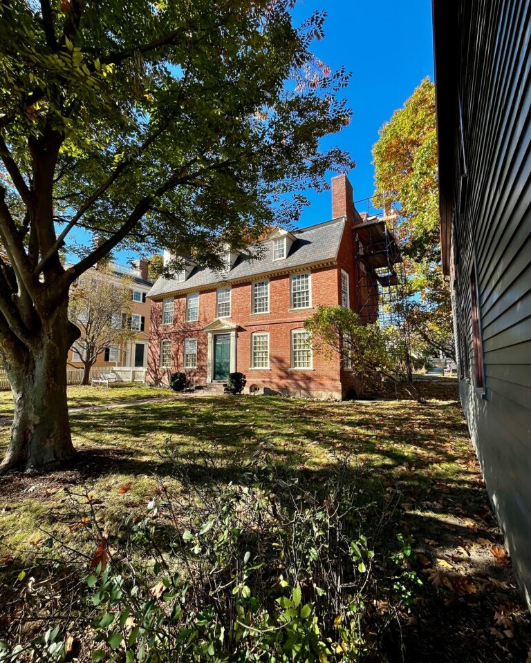 The Derby House in Salem, Massachusetts, viewed from a tree-lined yard, showcasing its red brick Georgian architecture, central green door, and surrounding historic buildings under a clear blue sky.
