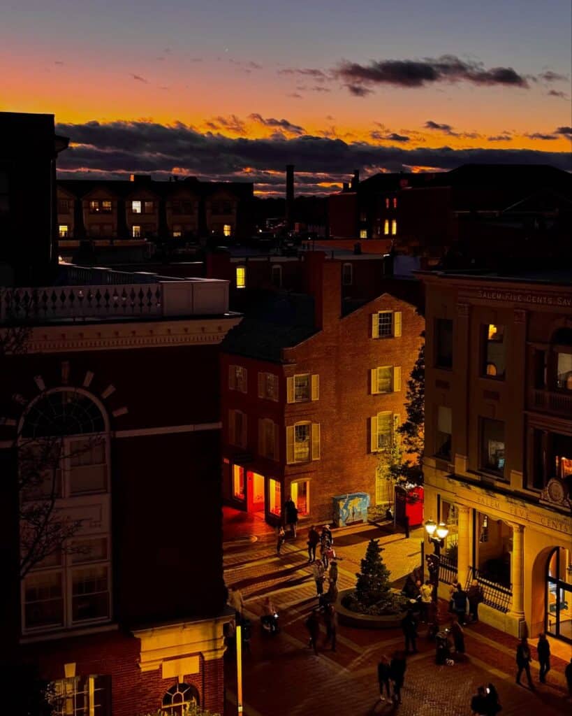 Downtown Salem with buildings illuminated against the backdrop of a vibrant sunset, showcasing colorful orange and blue hues in the sky while the street lights below bring the city to life.