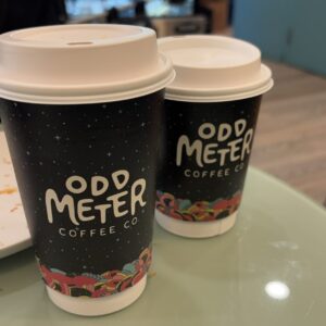 Two black coffee cups with white lids from Odd Meter Coffee Co. sit on a glass table. The cups feature a starry background and colorful abstract designs at the bottom.