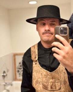 : A person wearing a tan overall with a mushroom pattern, a black shirt, and a black wide-brimmed hat takes a selfie in a well-lit bathroom with neutral decor.