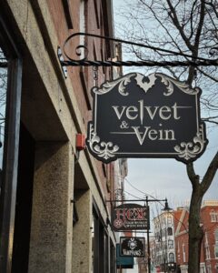Signs for Velvet & Vein, Hex Old World Witchery, and Vampfangs line a street in Salem, Massachusetts, with historic brick buildings and leafless trees in the background.