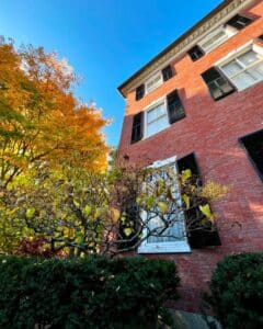 Close-up of the Andrew-Safford House in Salem Massachusetts, with its red brick façade, black shutters, and golden autumn leaves in the foreground.