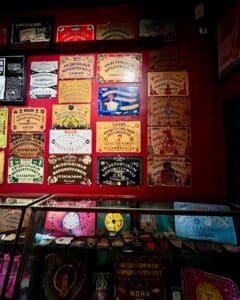 A colorful wall display at the Salem Witch Board Museum in Salem, Massachusetts, featuring an array of vintage and modern Ouija boards with intricate designs. Below the wall, a glass display case holds additional mystical artifacts and unique planchettes.