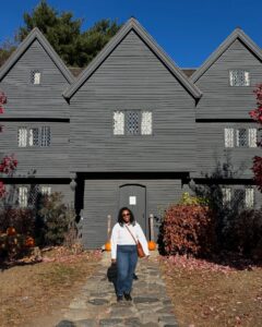 A woman stands in front of The Witch House in Salem, Massachusetts. The dark, historic building has steep gable roofs and diamond-paned windows, with pumpkins decorating the entrance pathway.