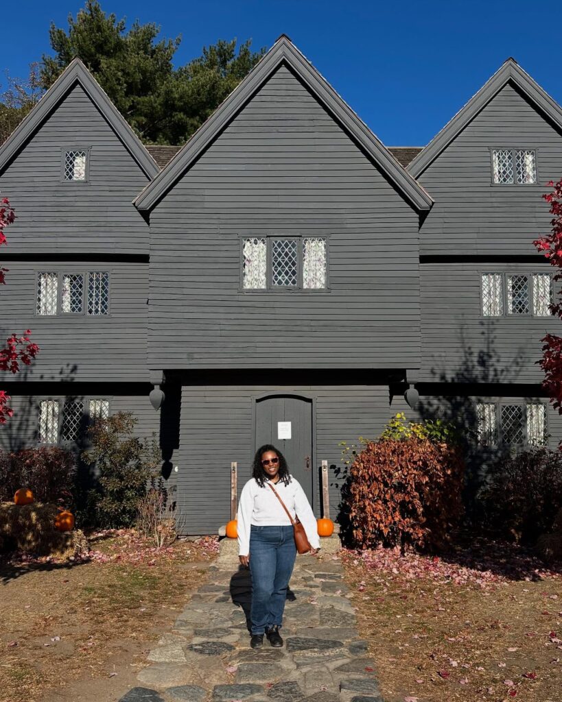 A woman stands in front of The Witch House in Salem, Massachusetts. The dark, historic building has steep gable roofs and diamond-paned windows, with pumpkins decorating the entrance pathway.