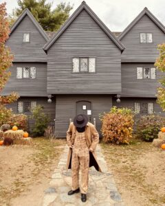A person dressed in a tan coat, patterned overalls, and a black hat stands on a stone pathway leading to the Salem Witch House, a black, gabled 17th-century building surrounded by autumnal foliage and pumpkins.