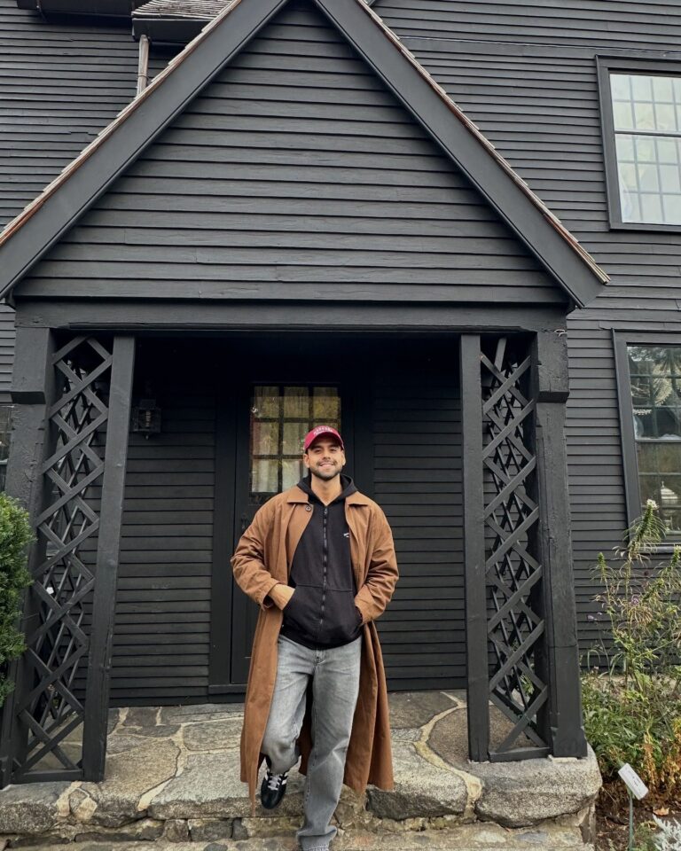 A man wearing a long brown coat and red cap stands on the stone steps in front of the House of the Seven Gables in Salem, Massachusetts. The dark wooden exterior and lattice details frame the entrance behind him.