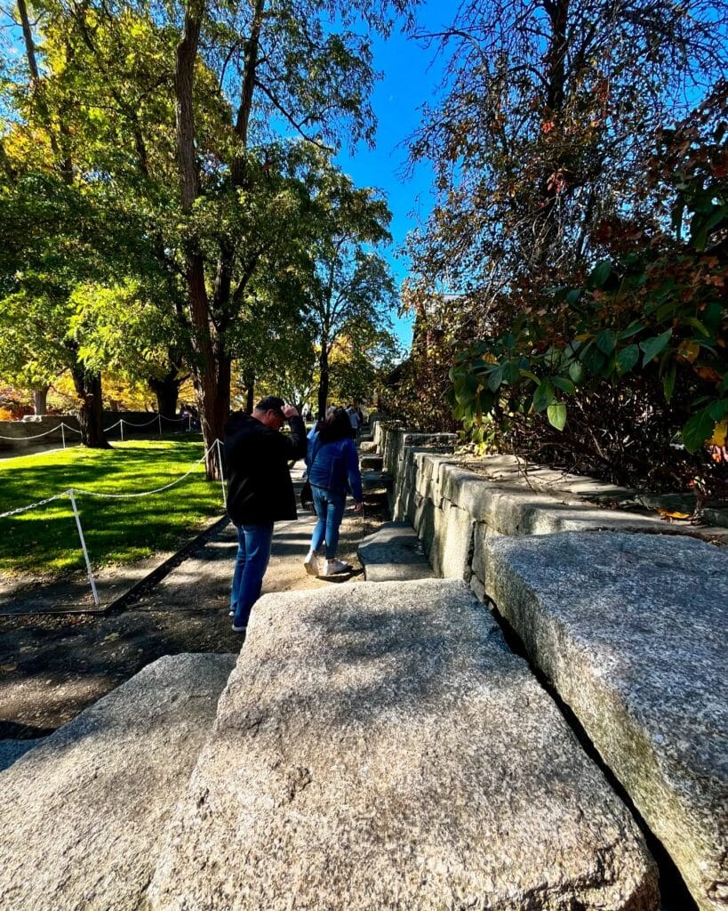 People walking through the Salem Witch Trials Memorial in Salem, Massachusetts, surrounded by large stone slabs engraved with victims' names. The area is shaded by trees with bright green foliage under a clear blue sky.