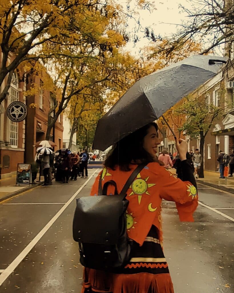 Woman holding an umbrella walks along Essex Street in Salem, Massachusetts during a rainy autumn day. The street is lined with vibrant fall foliage, and the woman wears a bright orange jacket decorated with sun and moon designs, carrying a black backpack.