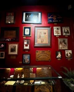 A vibrant red wall at the Salem Witch Board Museum in Salem, Massachusetts, adorned with framed vintage Ouija board advertisements, historical posters, and antique photographs above a display case of Ouija boards and planchettes.