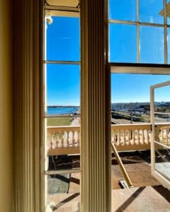 A view of the Salem waterfront seen through the tall windows of the Salem Custom House in Salem, Massachusetts, with bright blue skies, a balcony railing, and a scenic harbor in the background.