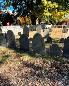 A row of weathered gravestones standing on grass covered with autumn leaves at Old Burying Point Cemetery in Salem Massachusetts, with a stone wall and trees adorned in fall colors in the background.