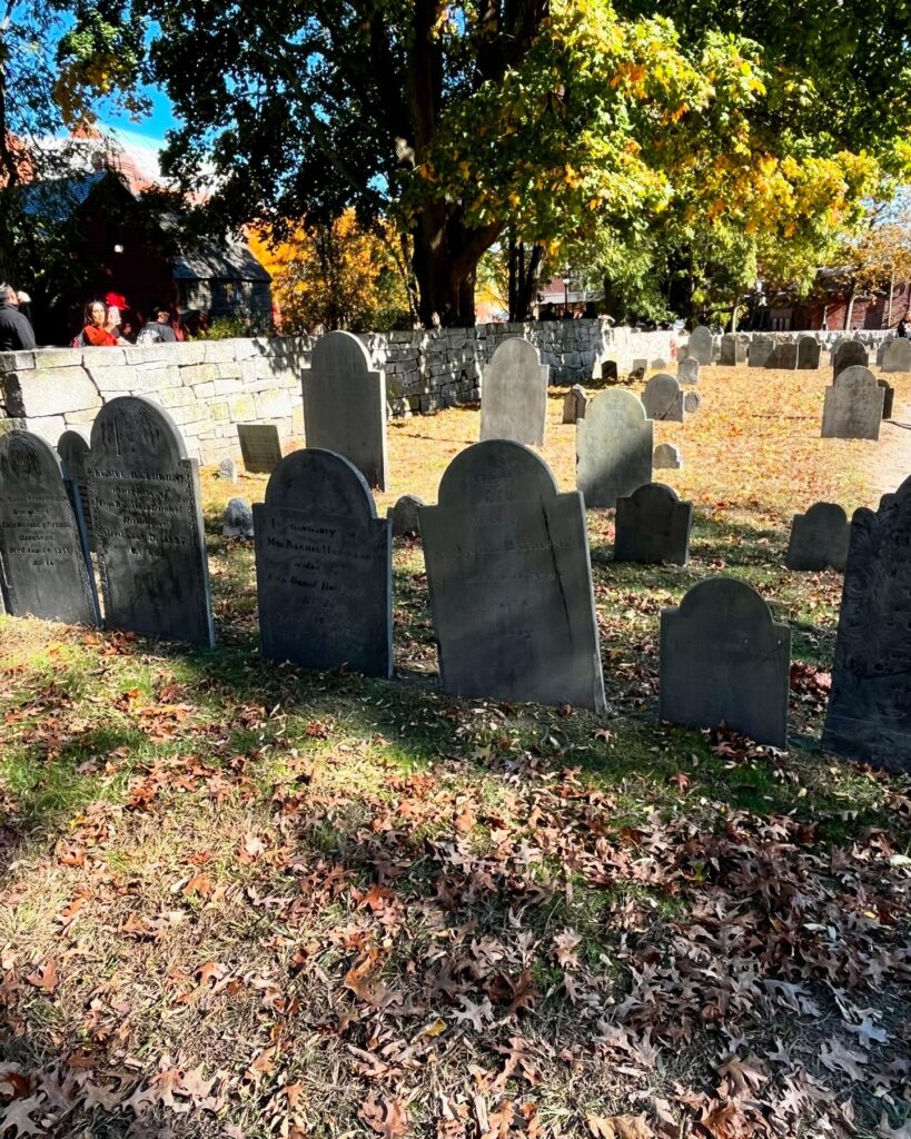 A row of weathered gravestones standing on grass covered with autumn leaves at Old Burying Point Cemetery in Salem Massachusetts, with a stone wall and trees adorned in fall colors in the background.