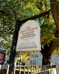 A sign for the First Church in Salem, Unitarian Universalist, founded in 1629, hanging near a wooden fence under a tree with green and yellow leaves, with the church and nearby buildings visible in the background.