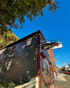 The West India Goods Store in Salem, Massachusetts, featuring dark green wooden siding, a hanging gold sign, and a bright blue sky with trees framing the historic building.
