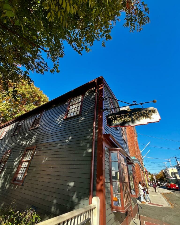The West India Goods Store in Salem, Massachusetts, featuring dark green wooden siding, a hanging gold sign, and a bright blue sky with trees framing the historic building.