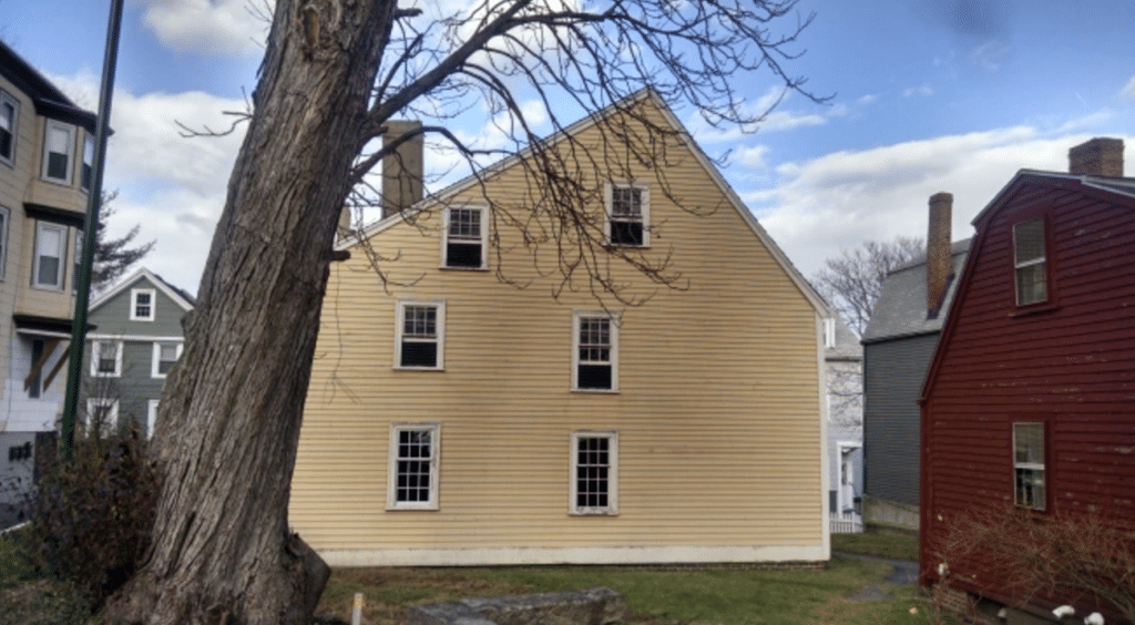 This image shows the rear view of the Gedney House in Salem, Massachusetts, emphasizing its simple yet historic First Period architecture. Built in 1665, the house features a steeply pitched roof and a yellow clapboard exterior. A large tree in the foreground adds natural character, while neighboring colonial-style buildings, including the red structure to the right, provide a contrast to the Gedney House's appearance. The open lawn and preserved surroundings offer a glimpse into the property’s historical context, making it a key part of Salem’s colonial heritage.