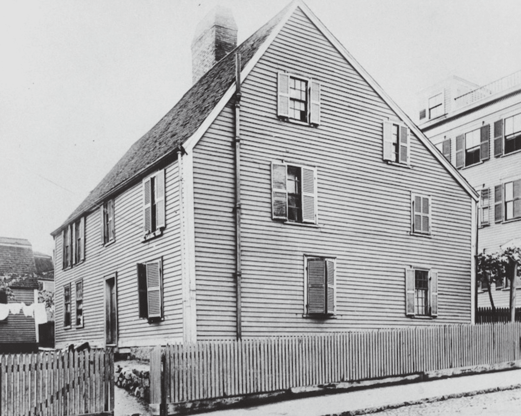 This historic photograph captures the Gedney House in Salem, Massachusetts, during the 19th or early 20th century. The colonial-style clapboard exterior and steeply pitched roof remain prominent features, reflecting its original 17th-century construction. The shutters on the windows and the picket fence enclosing the property highlight modifications made during its later use as a boarding house. The surrounding urban environment, visible in the background, shows the evolving neighborhood as Salem grew from a colonial settlement into a bustling city. This image showcases the house’s enduring presence through centuries of change.