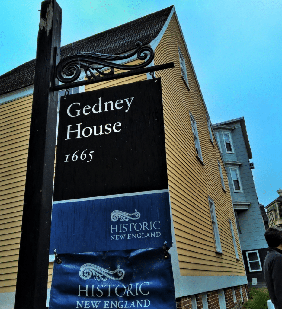 This close-up image highlights the signage for the Gedney House, a historic landmark in Salem, Massachusetts, built in 1665. The sign prominently displays the name "Gedney House" along with its construction date, underscoring its status as one of Salem's oldest surviving structures. Below, the logo of Historic New England is featured, representing the organization that preserves and manages the property. Behind the sign, the iconic yellow clapboard siding and steeply pitched roof of the house are visible, offering a glimpse of the First Period architecture that makes this home a significant