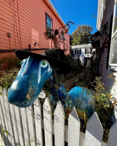A vibrant blue horse sculpture wearing a witch's hat stands behind a white picket fence, surrounded by Halloween decorations in a Salem, Massachusetts alley.