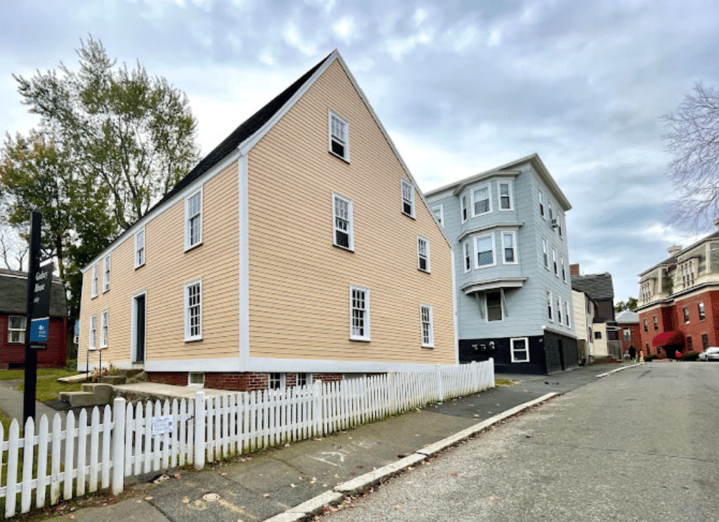 This modern photograph showcases the Gedney House, located on a quiet street in Salem, Massachusetts. Built in 1665, the house features a yellow clapboard exterior, steeply pitched roof, and white trim, all characteristic of First Period colonial architecture. The surrounding white picket fence adds to its historical charm, while nearby structures reflect the changing architectural landscape of the neighborhood. Managed by Historic New England, the Gedney House stands as a preserved landmark, offering visitors an authentic glimpse into Salem’s early history and its evolution over centuries.
