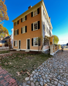 A wide view of the Hawkes House in Salem, Massachusetts, featuring peach clapboard siding, black shutters, white trim, and a cobblestone path leading to the entrance under a clear blue sky.