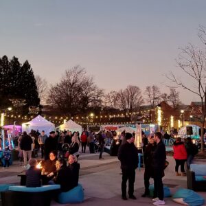 People gathering at the Salem Massachusetts Frozen Fire Festival, chatting by small fire pits and visiting vendor booths under soft evening lights.