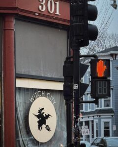 A storefront in Salem, Massachusetts, displays a sign reading "Witch City" with a silhouette of a witch on a broomstick. A traffic light and pedestrian crossing signal are in the foreground, with houses and street signs in the background.