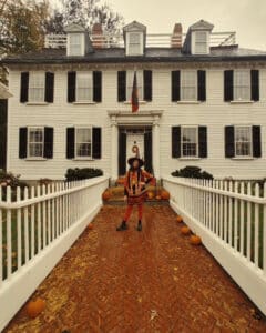 A woman in a witch costume stands in front of the Ropes Mansion, a historic white building with black shutters, decorated with pumpkins for Halloween in Salem, Massachusetts.