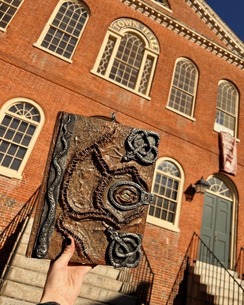 A hand holding the spellbook from Hocus Pocus in front of Salem's Old Town Hall. The book features an eye design with intricate stitches, while the brick façade and arched windows of the town hall are visible in the background.