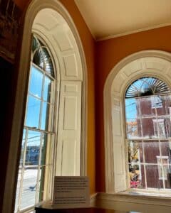 Interior view of the Salem Custom House in Salem, Massachusetts, featuring large arched windows with white trim, sunlight streaming through, and a historic desk replica with a placard describing its significance.