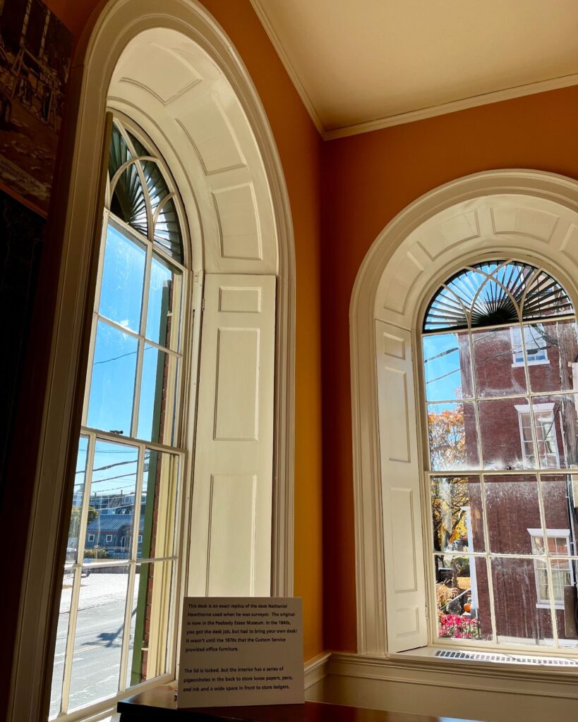 Interior view of the Salem Custom House in Salem, Massachusetts, featuring large arched windows with white trim, sunlight streaming through, and a historic desk replica with a placard describing its significance.