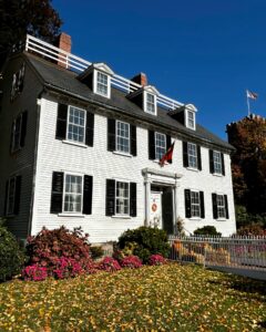 The Ropes Mansion in Salem, Massachusetts, a historic white colonial house with black shutters, surrounded by colorful autumn leaves, blooming flowers, and festive decorations under a clear blue sky.