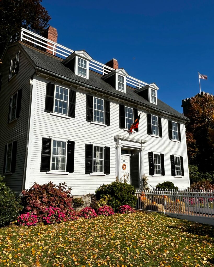 The Ropes Mansion in Salem, Massachusetts, a historic white colonial house with black shutters, surrounded by colorful autumn leaves, blooming flowers, and festive decorations under a clear blue sky.
