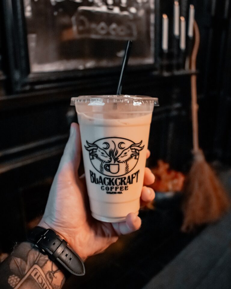 A hand holding a cup of iced coffee from BlackCraft Coffee in Salem, Massachusetts, with a spooky black interior and broom in the background.