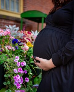 A close-up of a pregnant woman in a black dress gently holding her belly, surrounded by vibrant flowers in the Ropes Mansion Garden in Salem, Massachusetts.