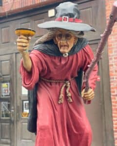 A detailed statue of an old witch in Salem, Massachusetts, wearing a red robe, a black pointed hat, and holding a staff and goblet, set against a backdrop of brick buildings.