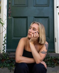 A young woman with blonde hair and tattoos sits thoughtfully on the steps of a historic green door in Salem, Massachusetts, surrounded by ivy.