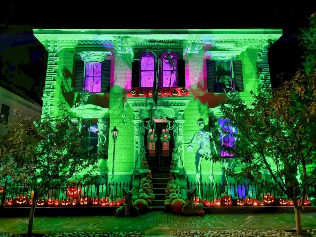 A vividly illuminated Victorian-style home in Salem, Massachusetts, glowing with green, purple, and orange lights, surrounded by Halloween decorations, skeletons, and carved pumpkins.